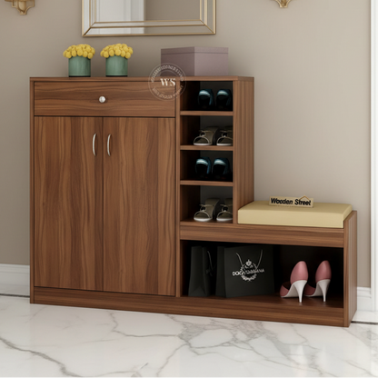 A brown wooden entryway shoe cabinet with a mirror, wall sconces, and a hallway in the background. The cabinet features a single drawer, two cabinet doors, open shoe storage shelves, a bench seat with a beige cushion, and a black lower compartment holding bags and shoes.