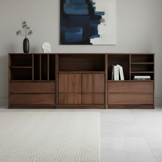 A modern wooden sideboard or console cabinet in a warm medium brown finish, styled in a minimalist living room setting with a beige sofa, a table lamp, a small potted plant, and a blue-gray abstract painting on the wall.
