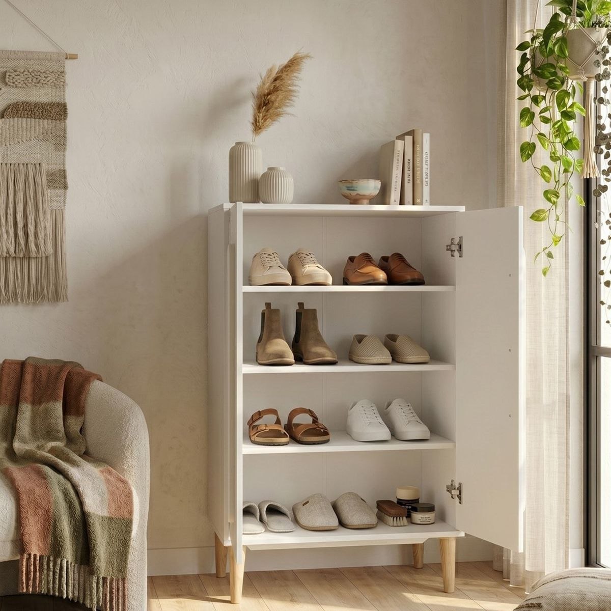 A white open shoe cabinet with wooden legs stands in a bright room, styled with various pairs of shoes on its shelves and decorative items on top. The cabinet is set against a warm beige wall next to a window with sheer curtains and a hanging green plant.
