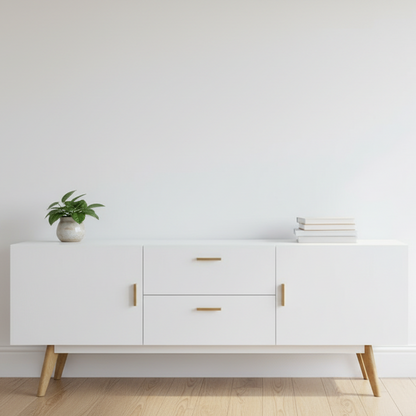A modern white sideboard or credenza with gold-tone handles and tapered wooden legs, styled in a contemporary room with two table lamps, a clock, books, and a decorative vase. Above it hangs a large abstract painting on a dark gray wall.