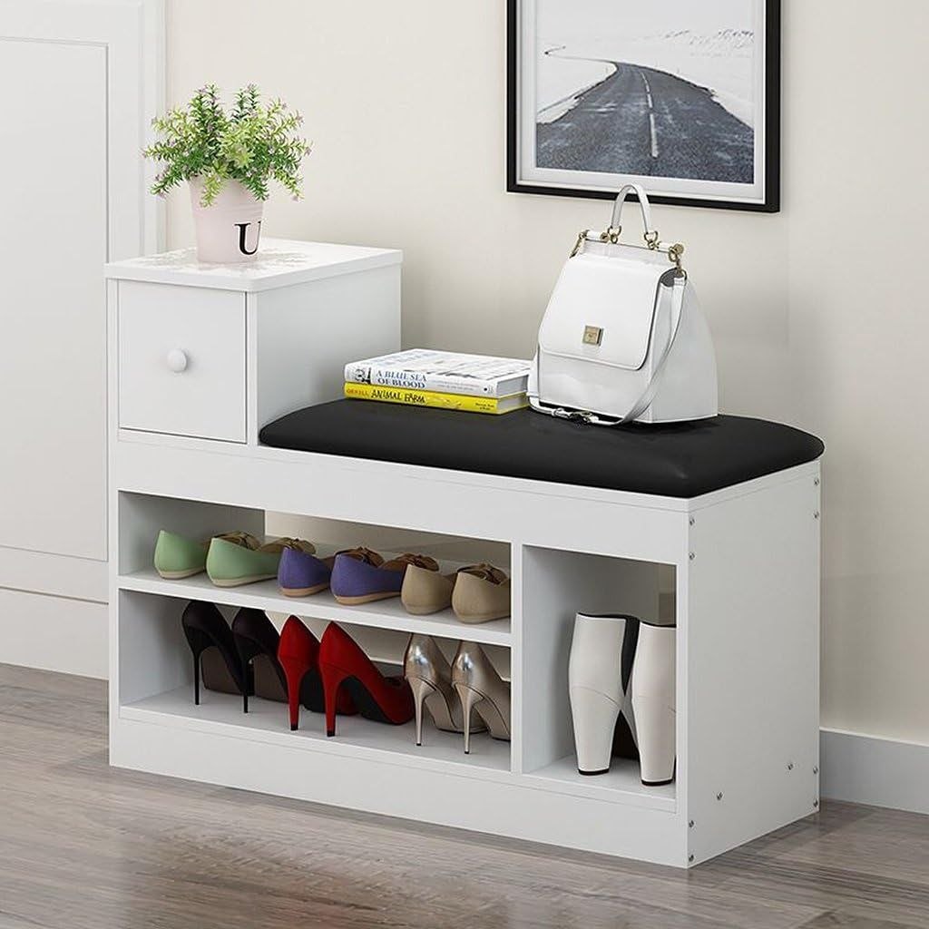 A white entryway shoe bench with a black cushioned seat, open storage shelves, and a small side cabinet. The bench is styled with several pairs of shoes, a white handbag, books, and a potted plant against a light beige wall.