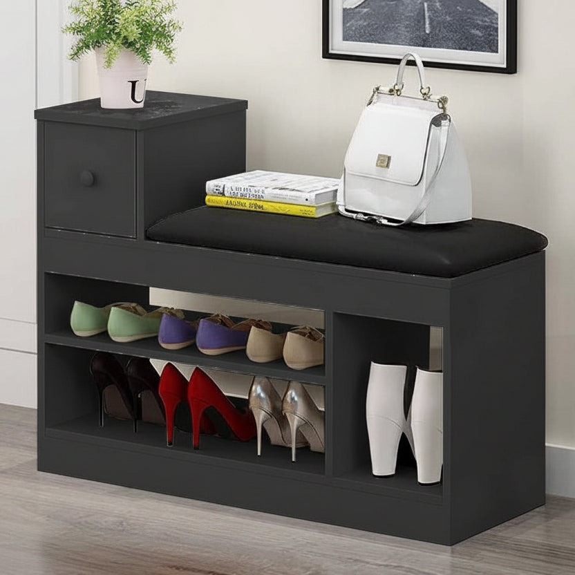 A dark gray entryway bench with a black cushioned seat, featuring a small side drawer, open shoe storage shelves, and display pockets. The bench is styled with a white handbag, books, a small potted plant, and several pairs of shoes, set in a home entryway environment.