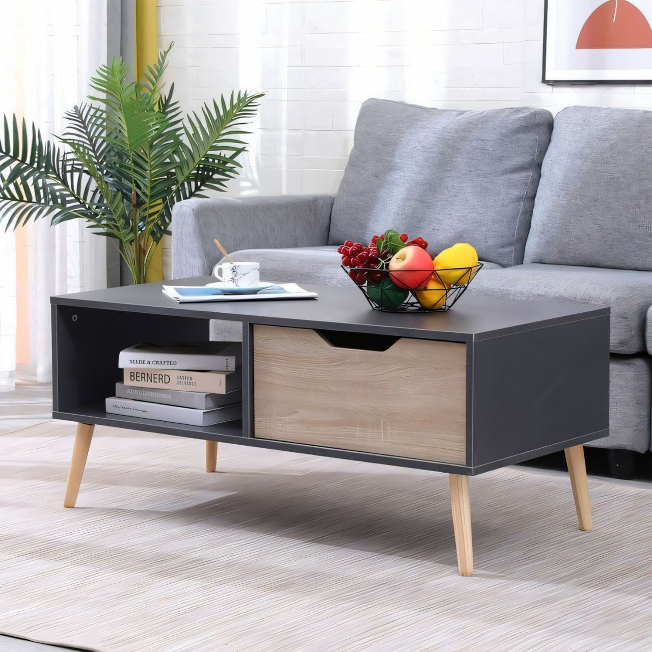 A modern low coffee table with a dark gray top and frame, light wood drawer front, and tapered light wood legs, styled in a living room setting with a gray sofa, fruit bowl, and books.