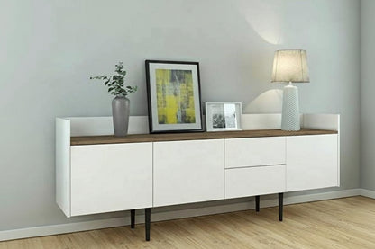 A modern white sideboard cabinet with a light brown wood-look top, black tapered legs, and decorative items including a vase, framed art, and a table lamp, set against a light gray wall and light wood floor.